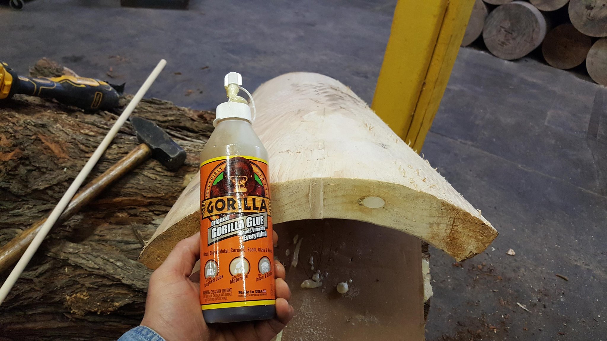 Gorilla Glue Original bottle held in front of the repaired IC WOOD half-log — finished logs stacked in background, IC WOOD production shop